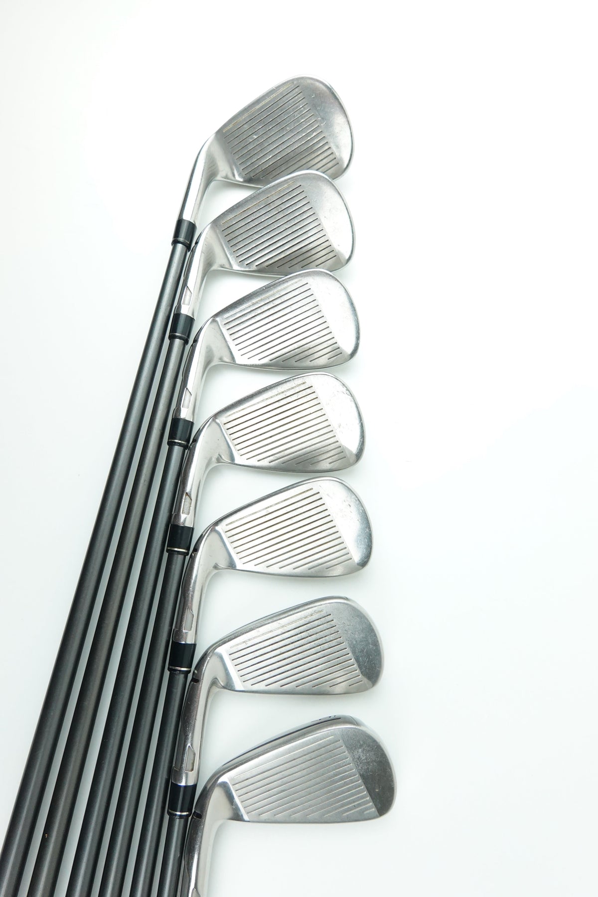 A set of eight golf iron clubs with silver metal heads and black shafts, arranged in a neat diagonal row on a clean white background.